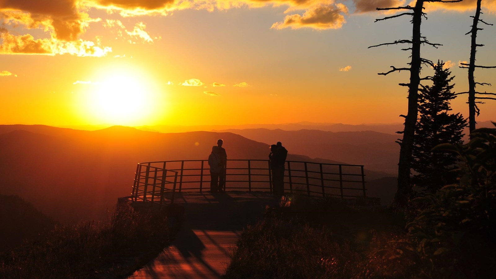 The sunsets silhouetting two people standing at a railing looking toward the sun. 