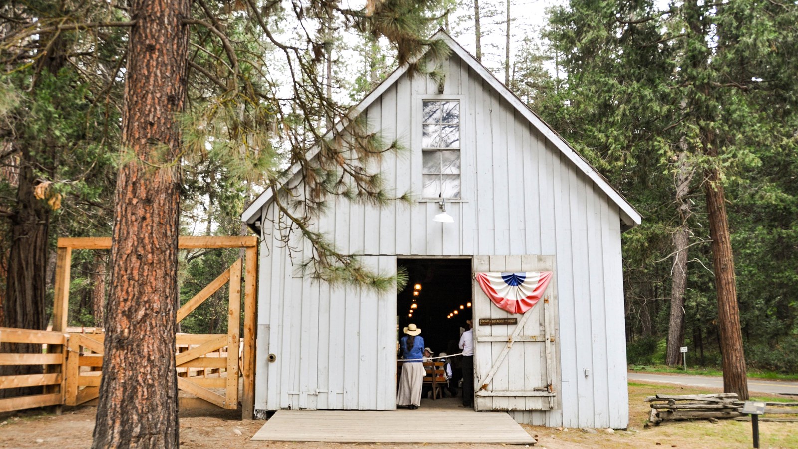A simple grey barn in a dirt area surrounded by trees
