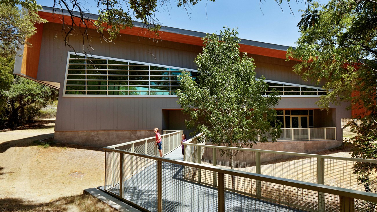 People stand on an elevated walkway leading to a large building with many windows and slanted roof.