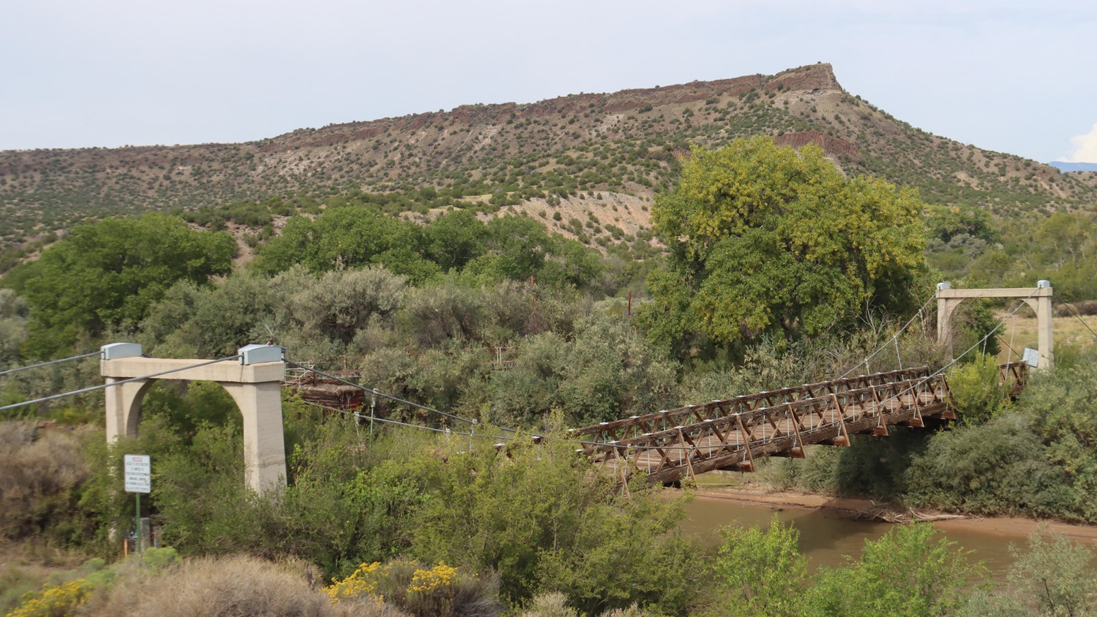 An old, rustic wooden bridge spans a muddy dirty river with a mesa visible in the background. 