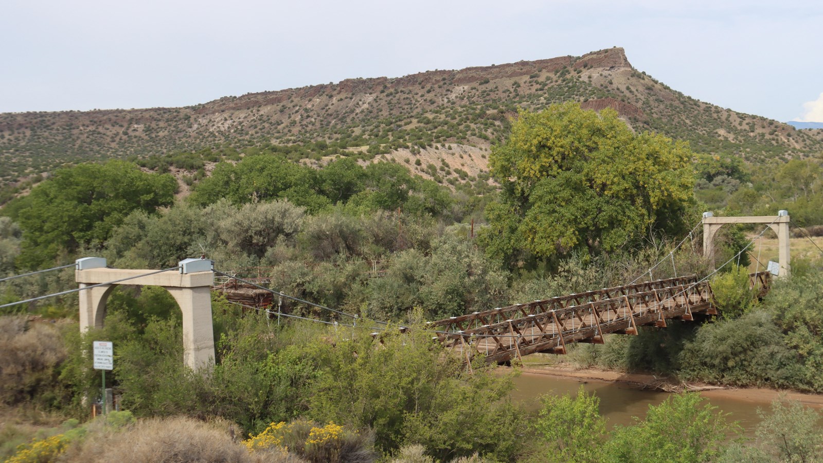 An old, rustic wooden bridge spans a muddy dirty river with a mesa visible in the background.