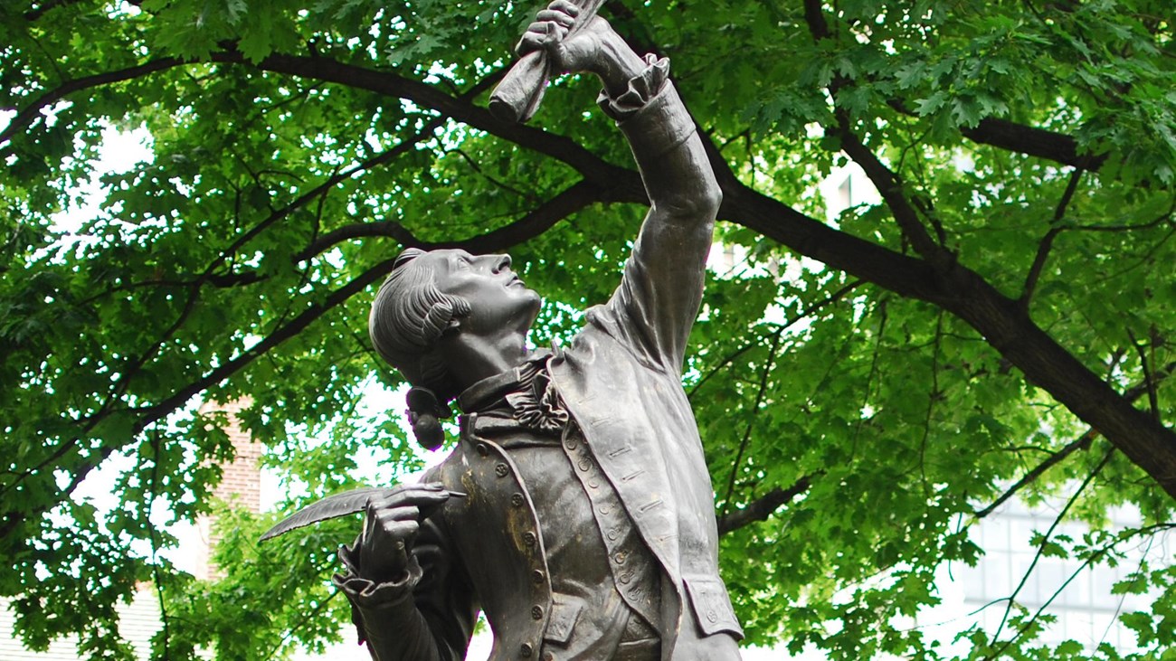 Color photo of a statue showing a man with quill pen and rolled scroll.