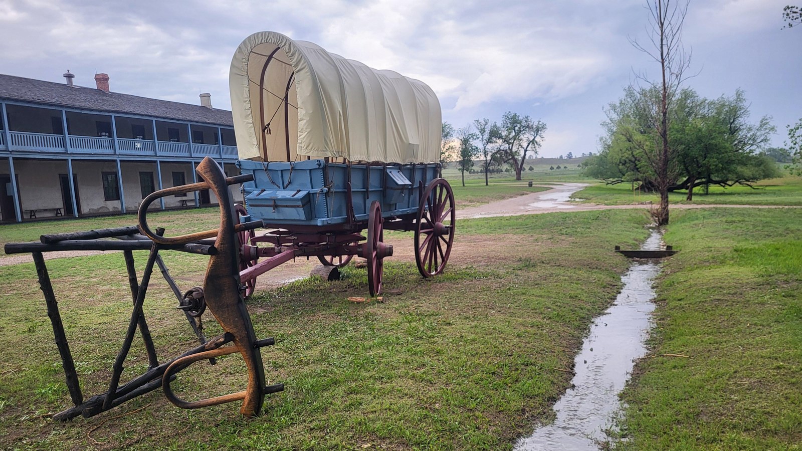 A historic covered wagon sits next to a small stream with a building behind it. 