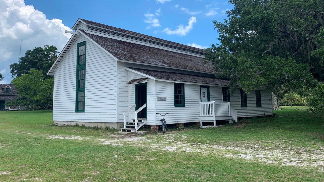 A white building with pitched roof in an open field with a large live oak to the right. 