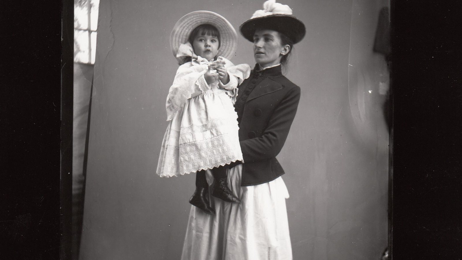 Woman in Victorian era attire, holding child in white gown.