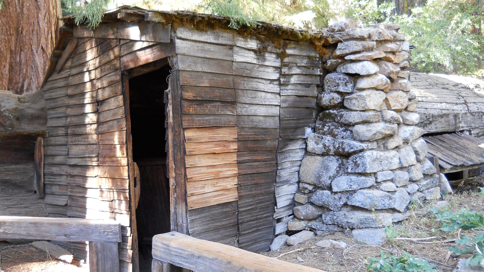A cabin with a stone chimney is built into a downed, giant sequoia log.