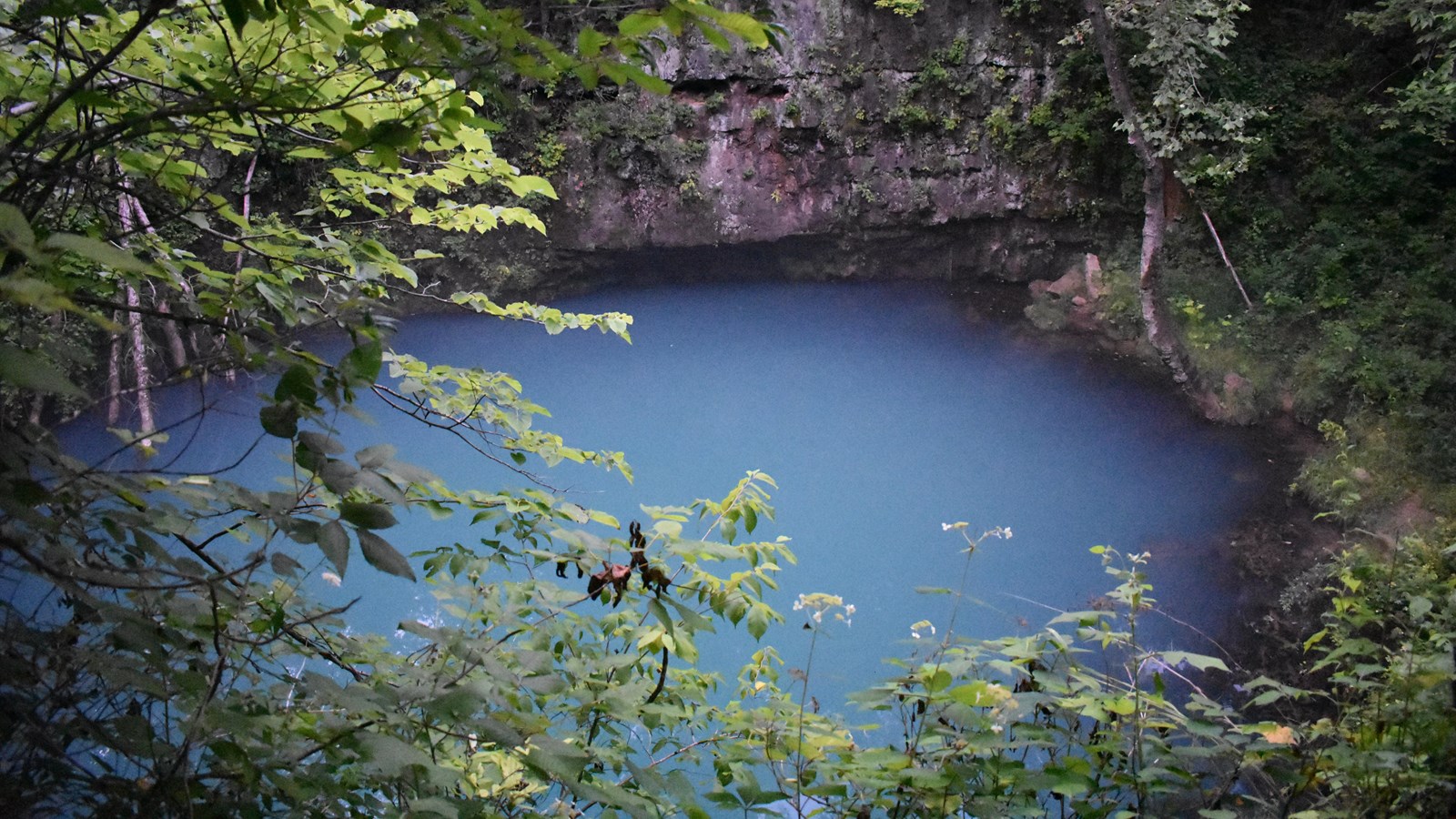 a blue round pool of water with grey rock ledge behind and green trees surrounding all sides