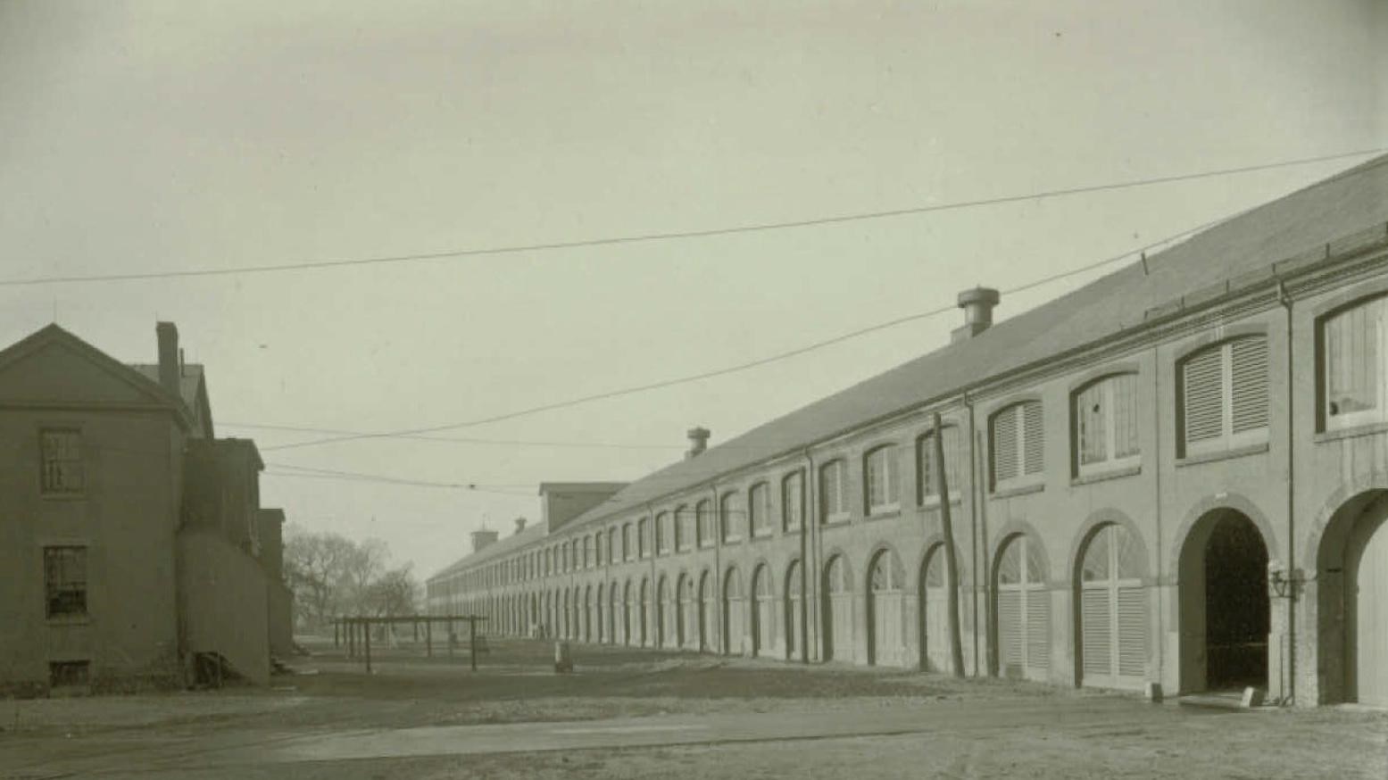 A black and white photo of a long building with multiple windows and a small house to the left.