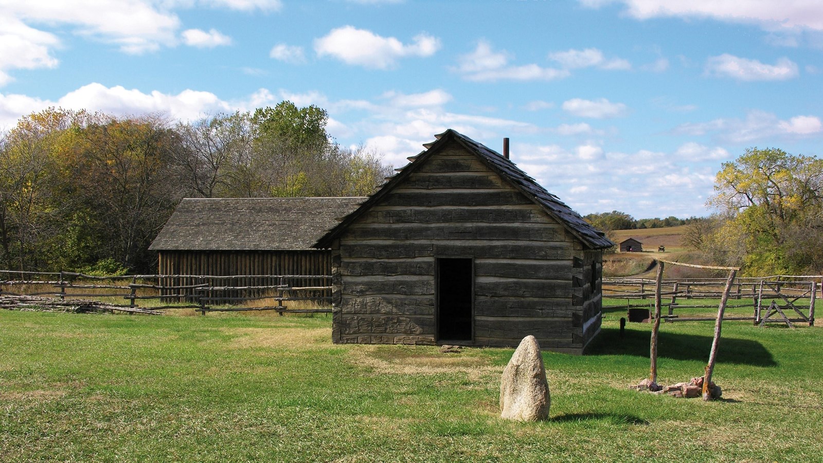 Two log cabin buildings with pitched roofs sitting in a grassy area with trees in the distance.