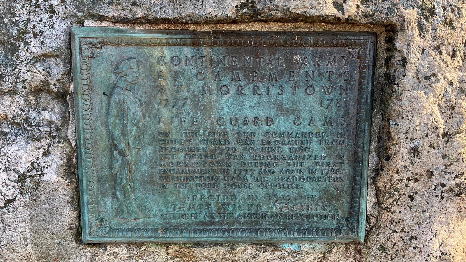A weathered metal plaque set into stone, with a Continental Soldier standing next to text.