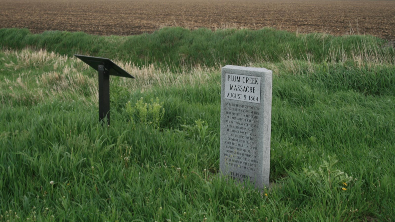 A rectangular, gray stone monument stands among prairie grass at the edge of a cultivated field. 