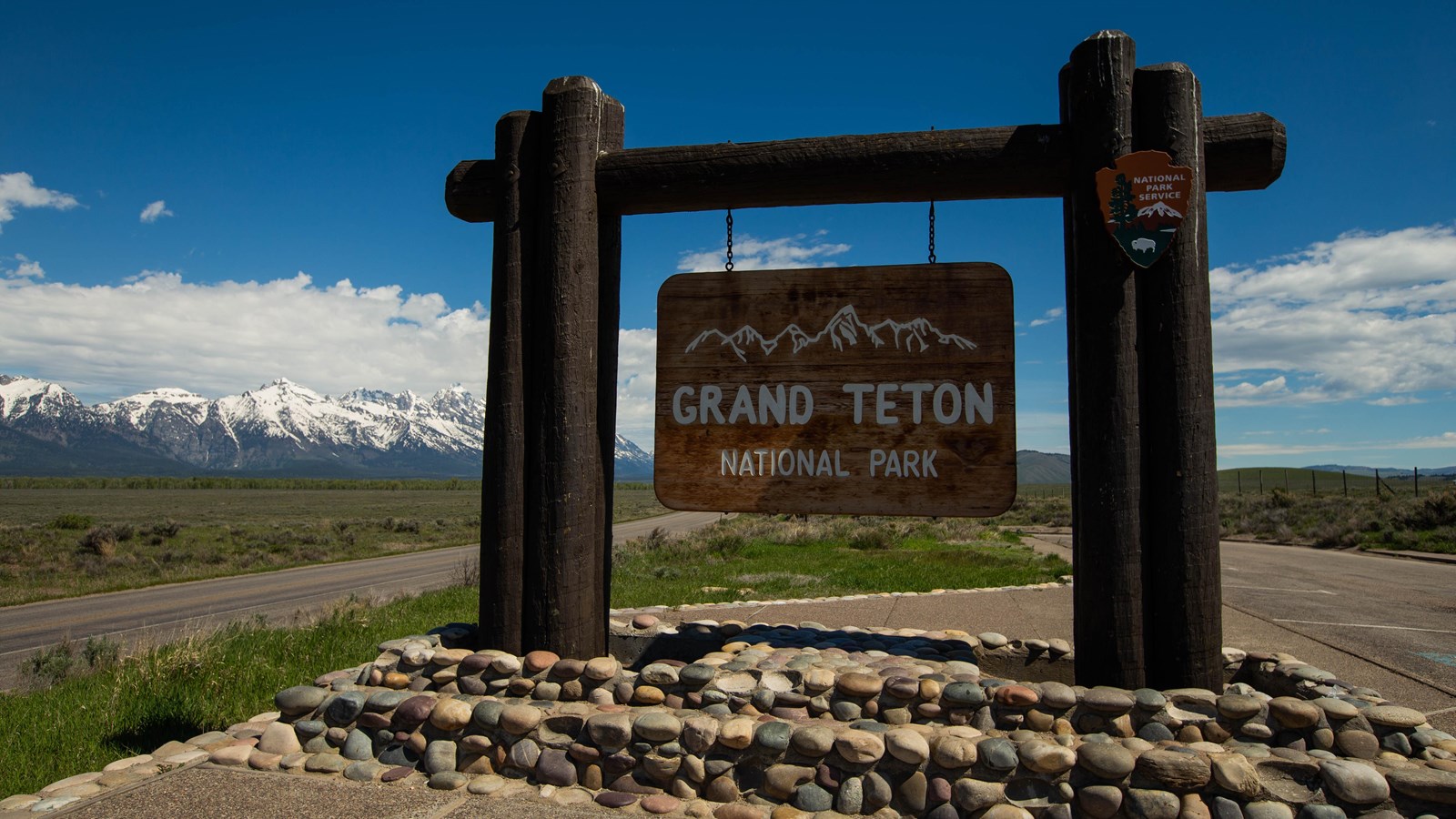 A sweeping view of the Teton Range and a large wooden sign that says Grand Teton National Park. 