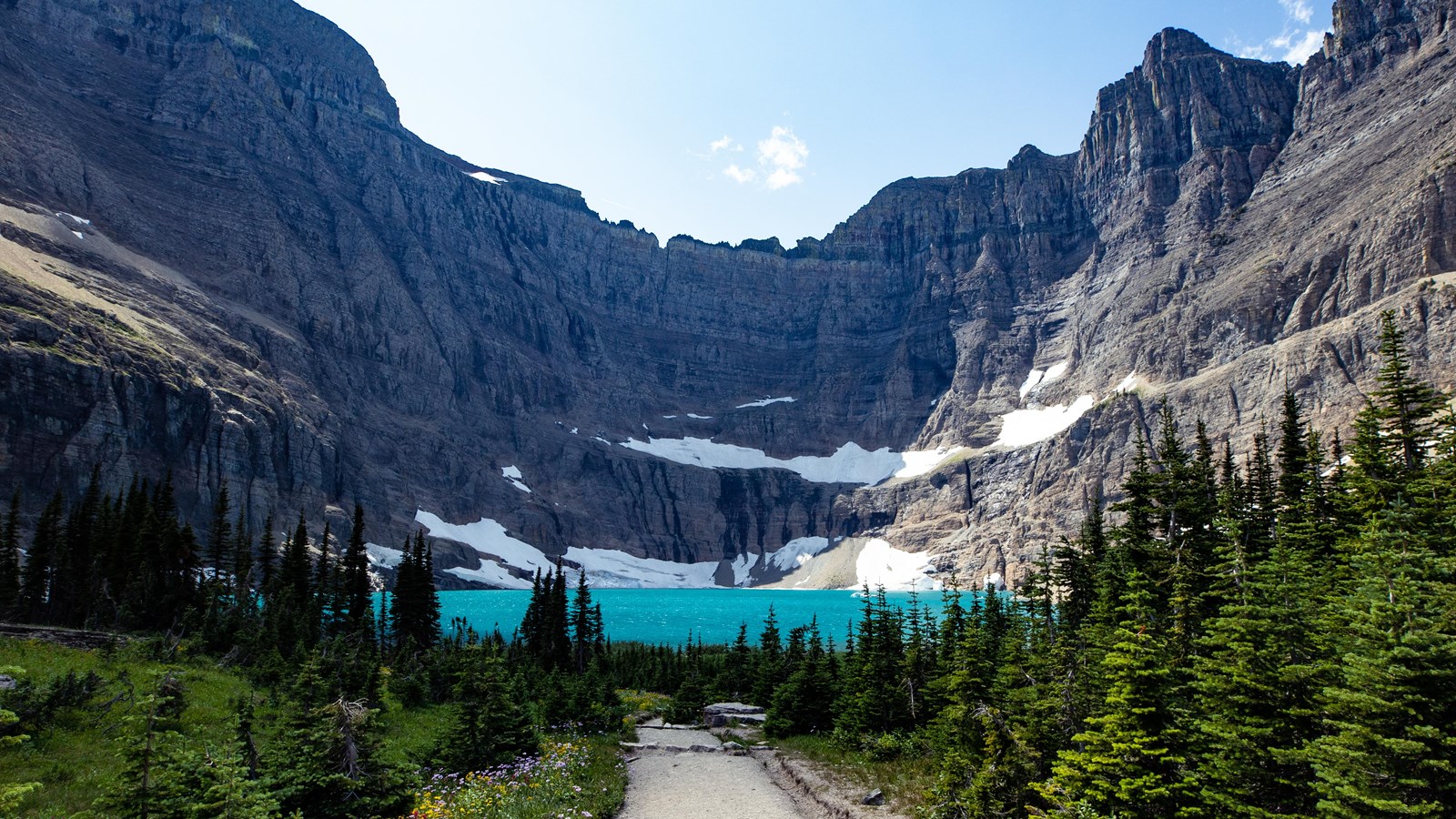 A bright blue lake sits under steep rocky cliffs with patches of snow. A dirt trail leads through th