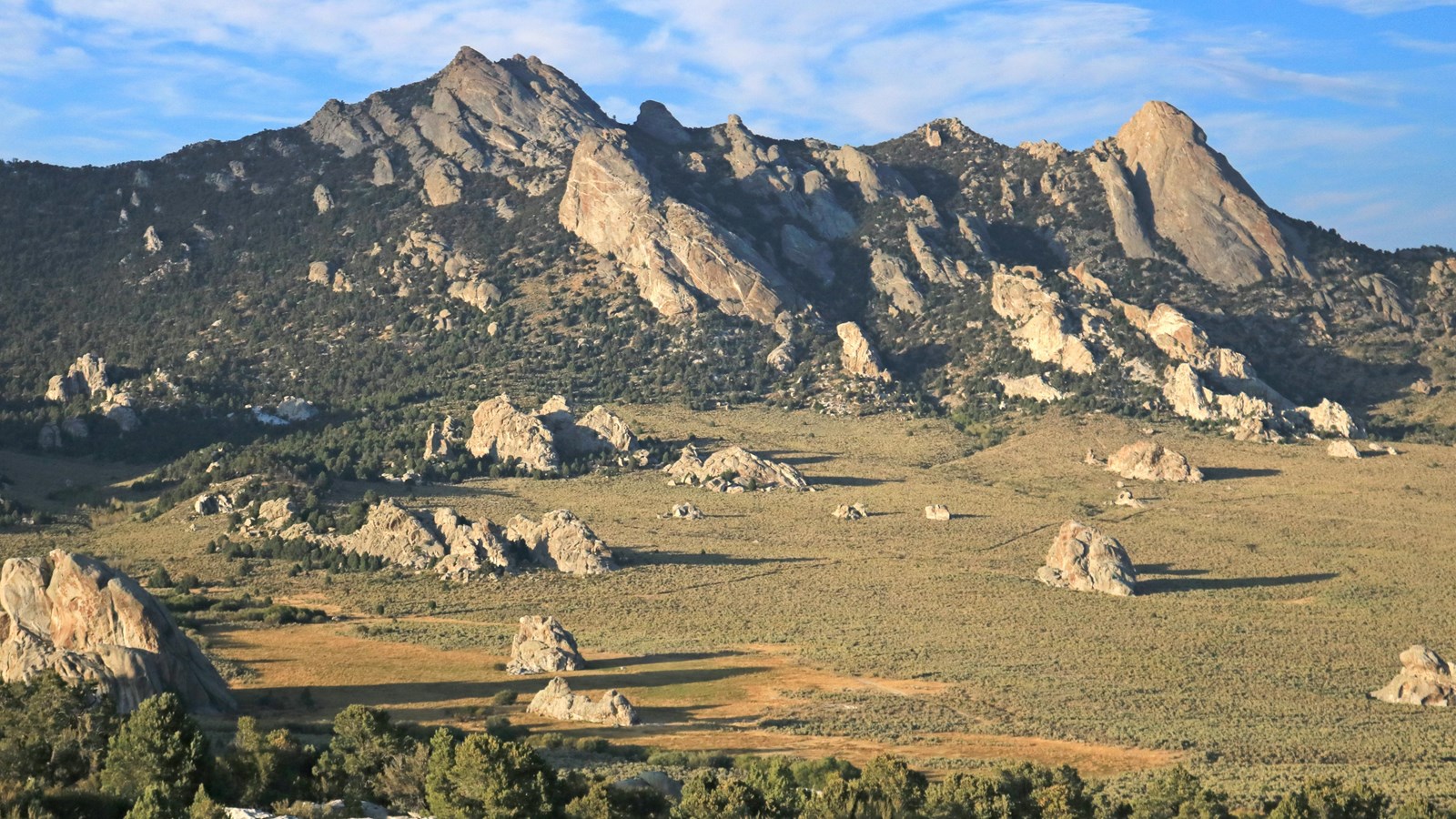Expansive view of a green meadow dotted with granite formations and craggy mountains in the distance