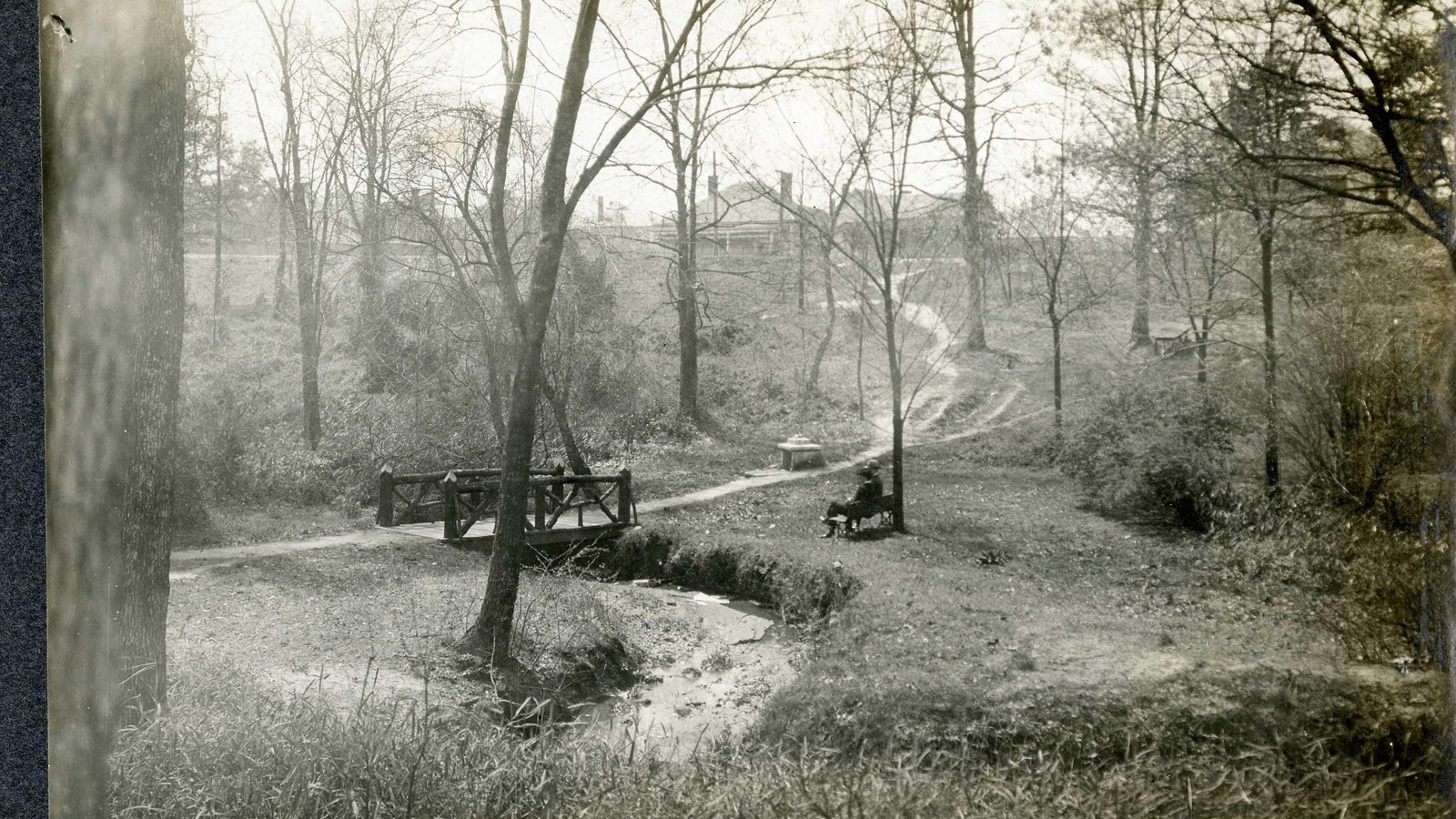 Black and white of dirt path over water with wooden bridge leading up hill with trees and bench