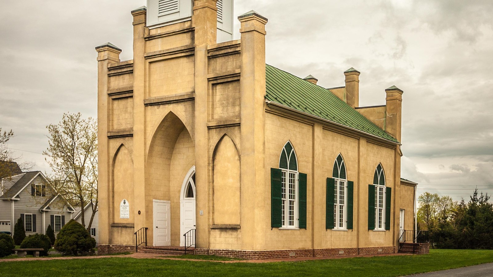 A small gothic revival style church has pointed arch windows and a buff color exterior.