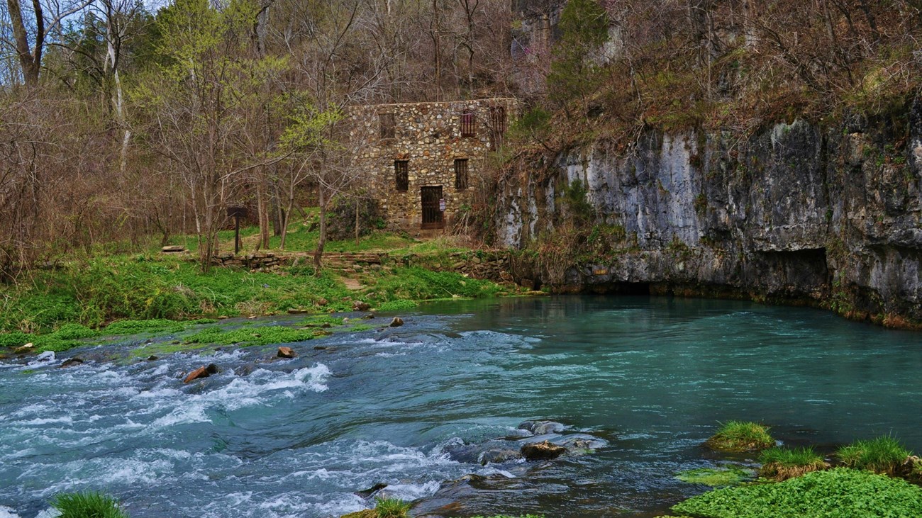 a rock building in distance with blue spring flowing in front, green vegetation around spring