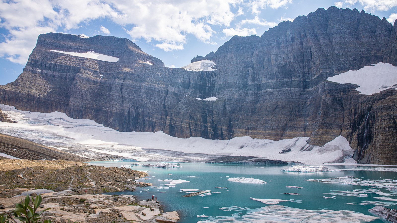 A turquoise lake with floating pieces of ice sits at the base of snow-speckled, striated cliffs.