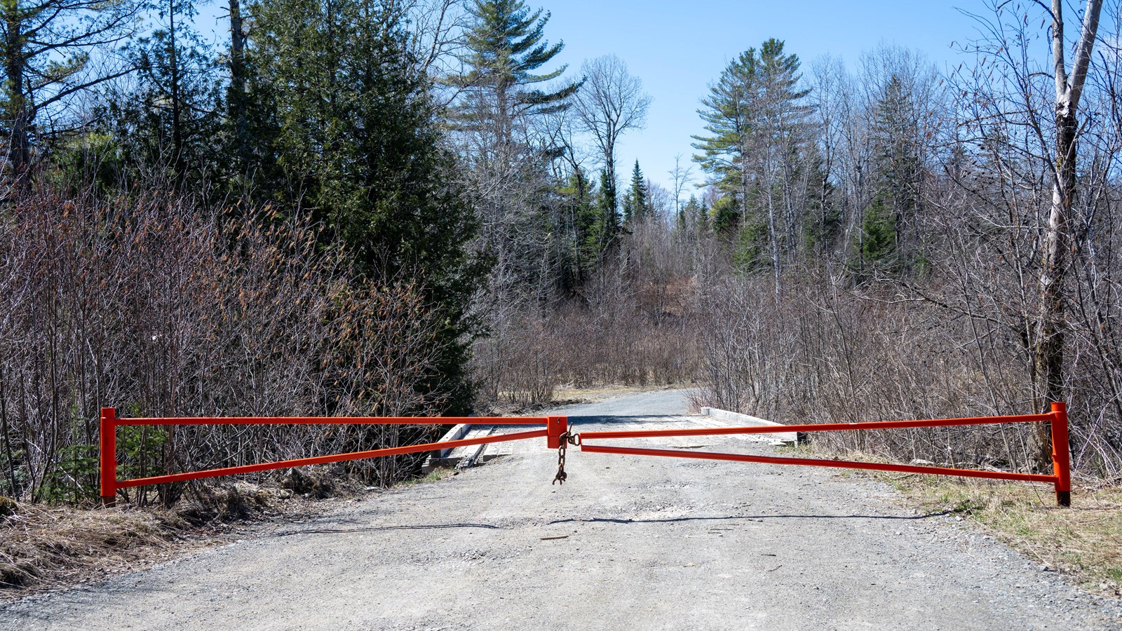 A large orange metal gate is locked closed across a gravel road in the woods.