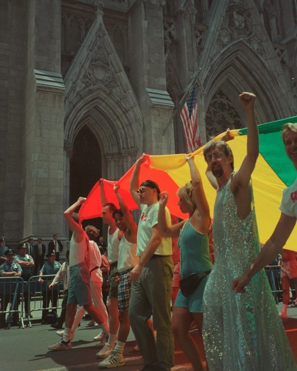 Gilbert Baker wears a sequin dress, holds a large Rainbow Flag up with other protestors
