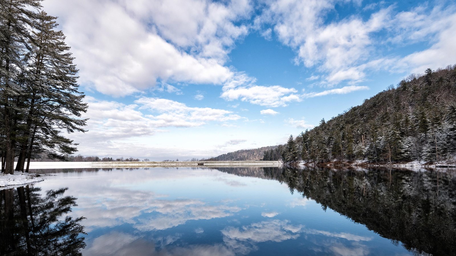 A winter scene of a lake framed by snowy trees on the left and a mountain ridge on the right.