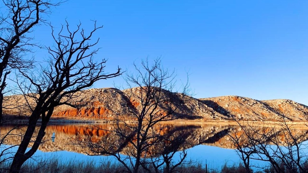 The water is glassy with mesas in the background.  There are trees along the shoreline. 