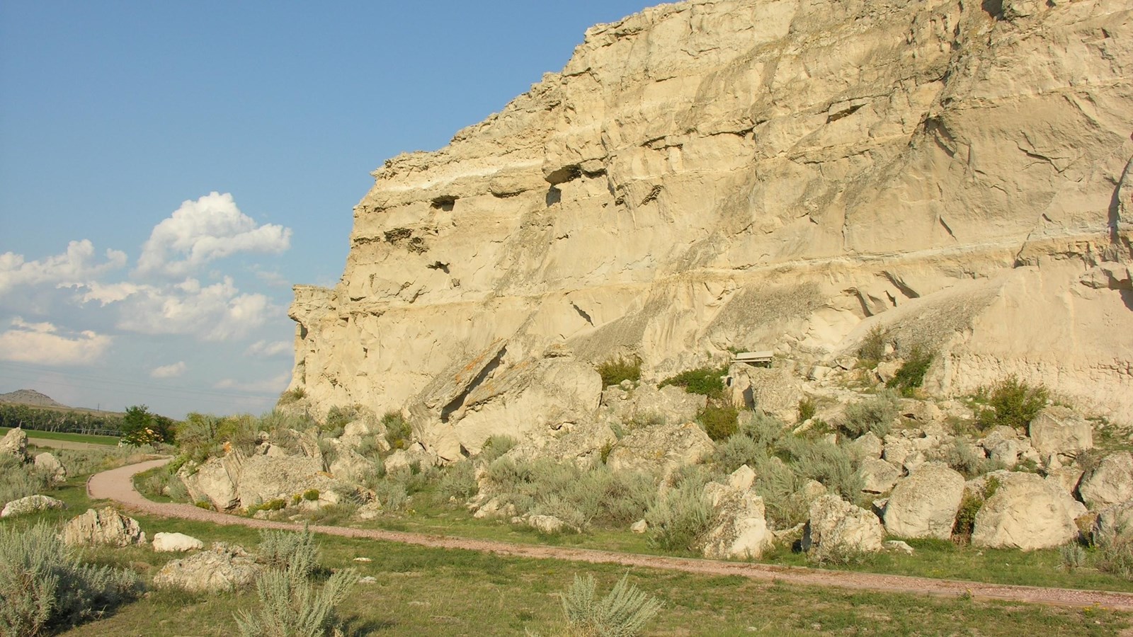 A path hugs close to a large sandstone bluff in a grassy area. Names are etched in the bluff.