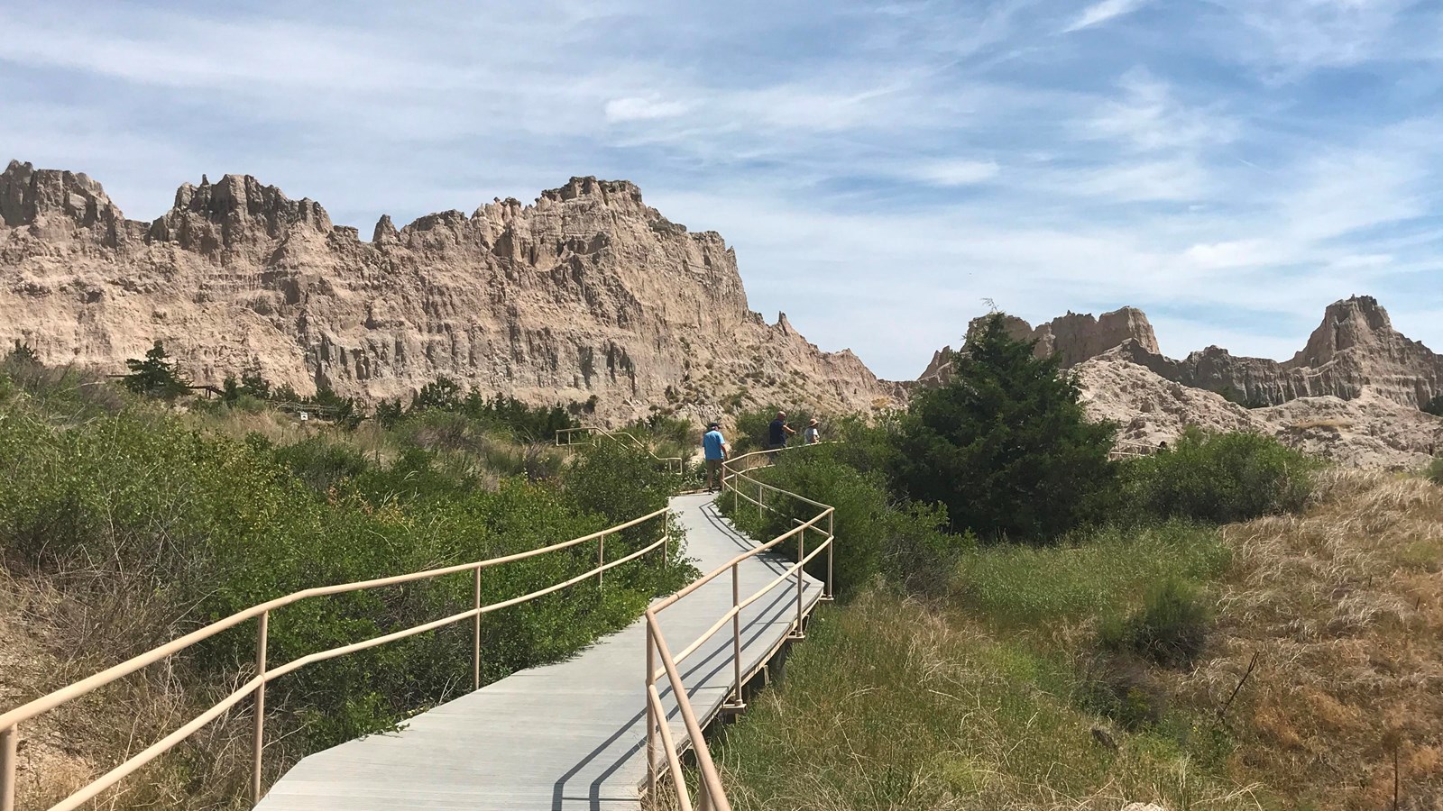 A sloped boardwalk proceeds through cedar trees and towards badlands buttes under a blue sky.