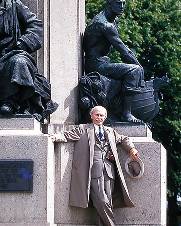 Two bronze statue of soldiers at rest in front stands a man in a tan suit holding a hat