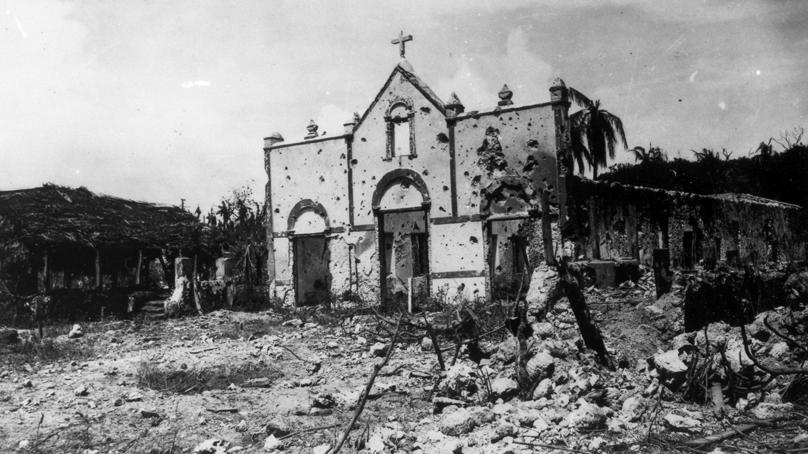 Black and white photo of the facade of a church full of bullet holes surrounded by ruins.