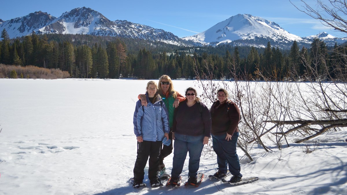 Four women in snowshoes standing on the edge of a snow-covered lake backed by volcanic peaks. 