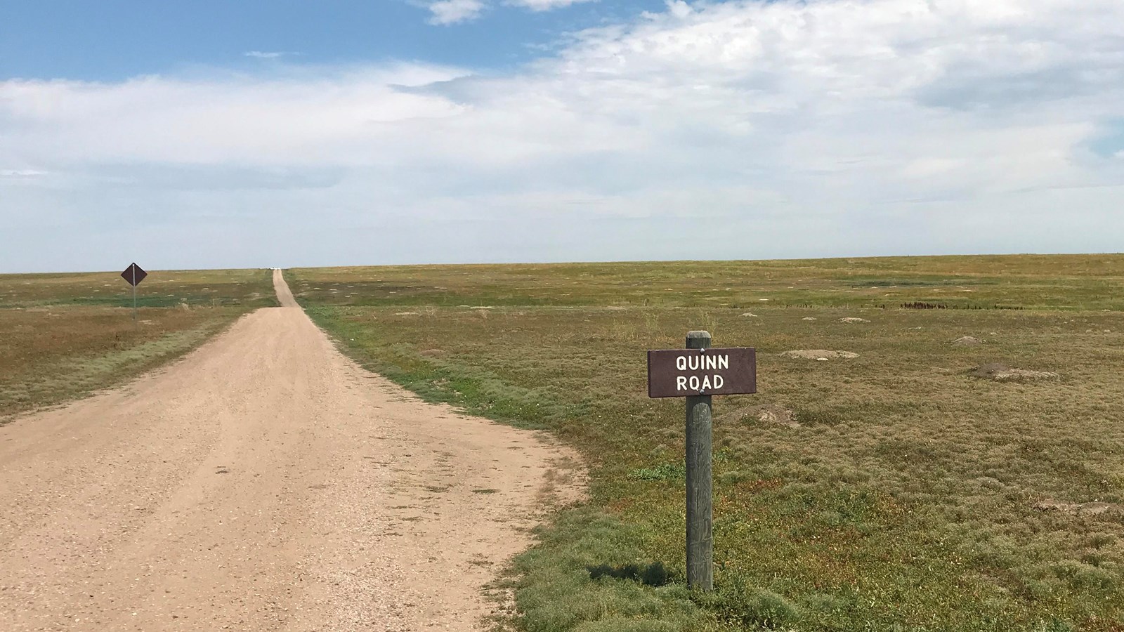 Winding dirt road alongside flat green grassy field under blue sky. A wooden sign reads 