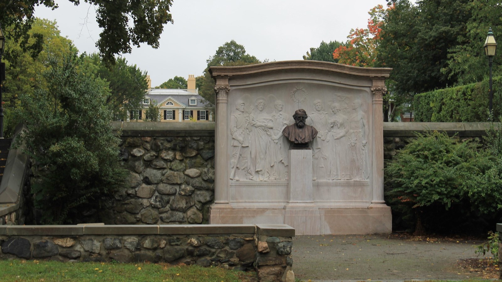 Monument with bas relief figures and central bust of Longfellow. Top of yellow mansion in background