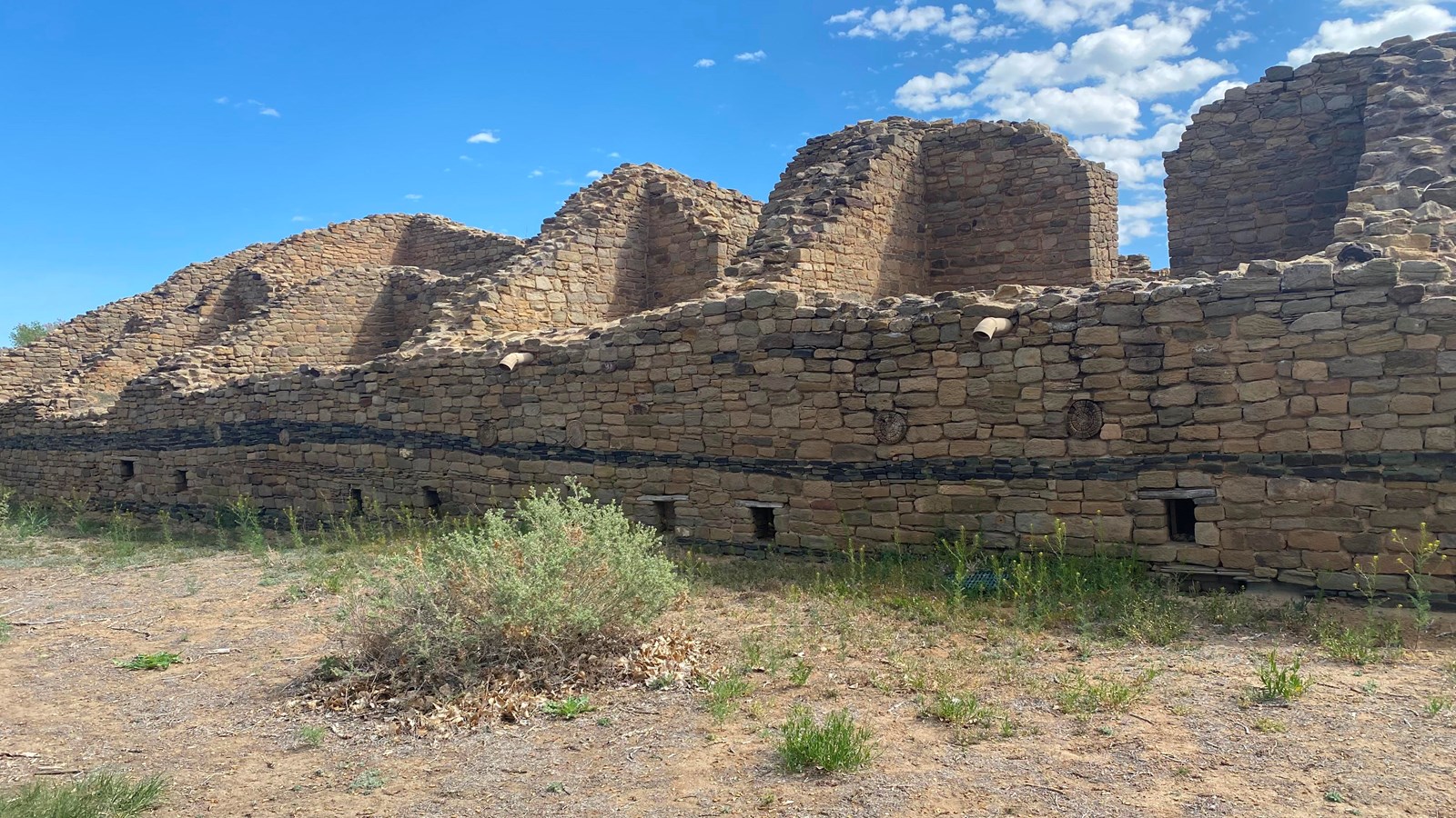 A long wall of stacked, tan sandstone blocks with a stipe of green rock in the middle.