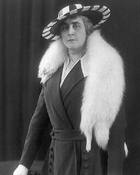 Woman standing wearing a hat and white fur stole.