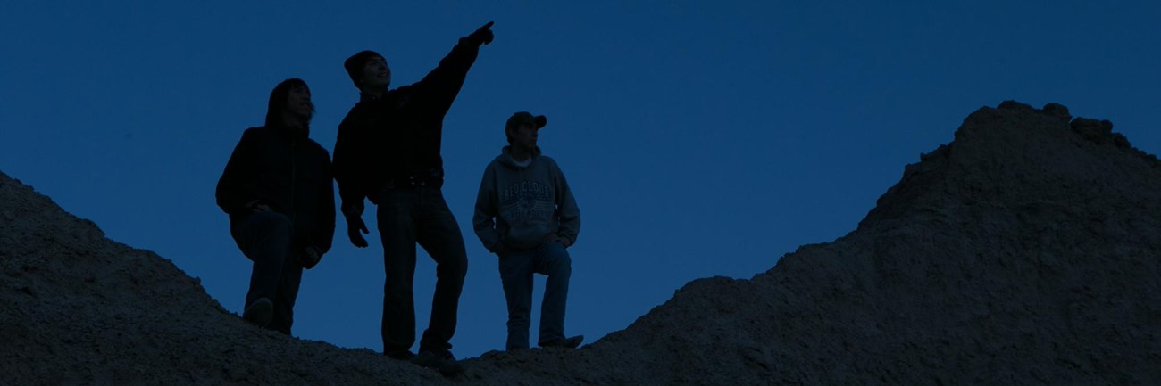 three people stand silhouetted atop a badlands butte, pointing at the night sky.