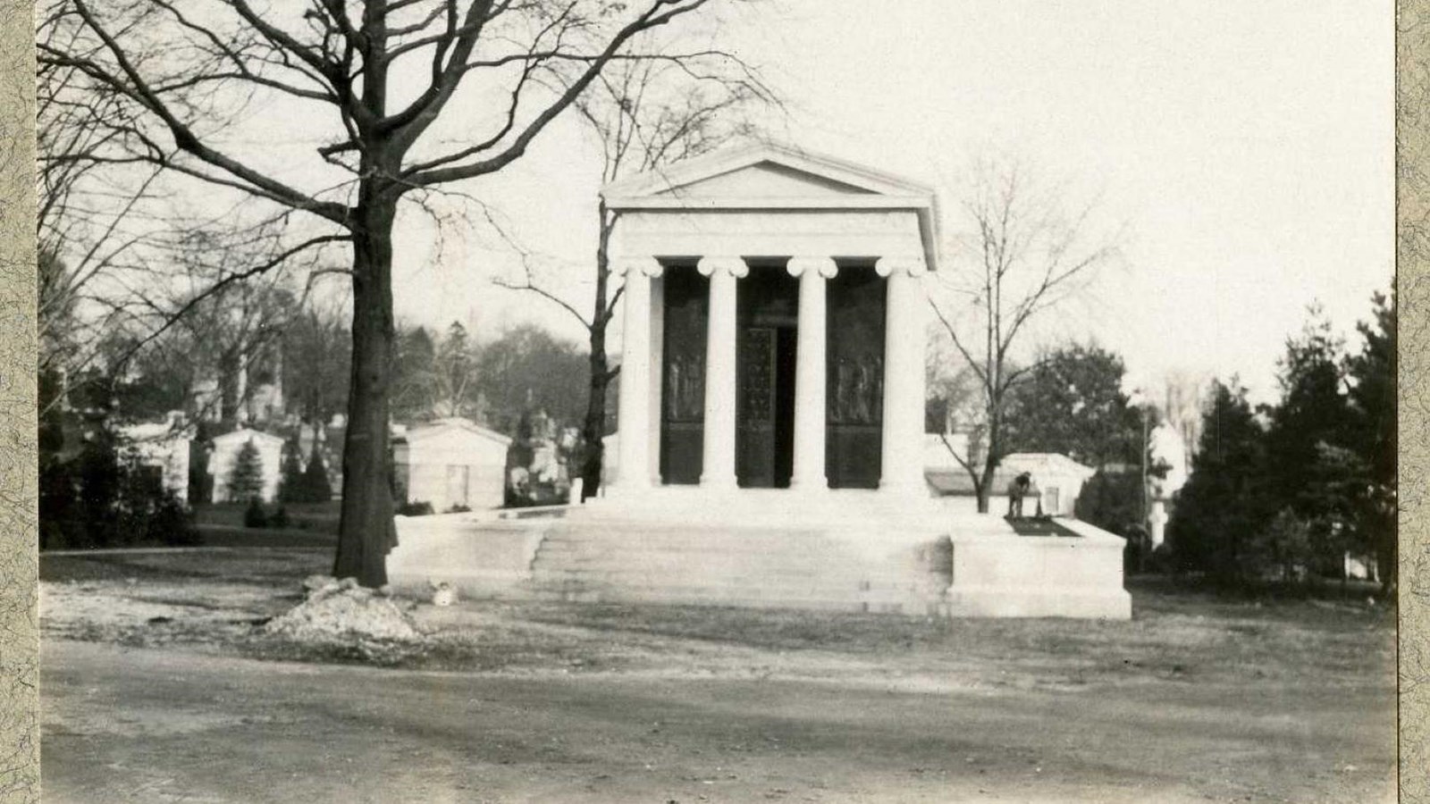 Black and white of white building with four columns, tree in front and behind, cemetery behind 