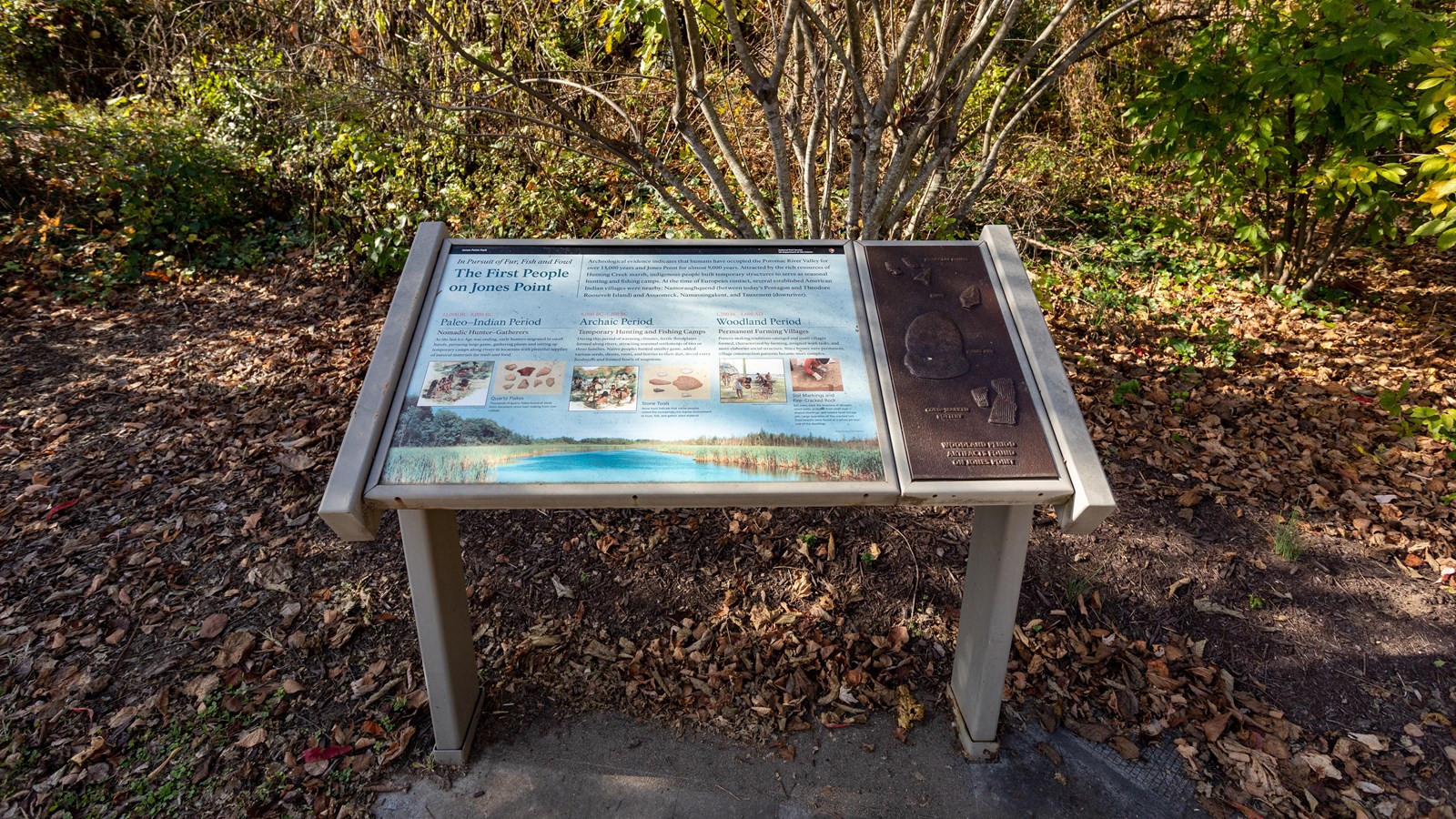 An angled exhibit panel faces a wooded area. The right side of the panel has tactile elements.
