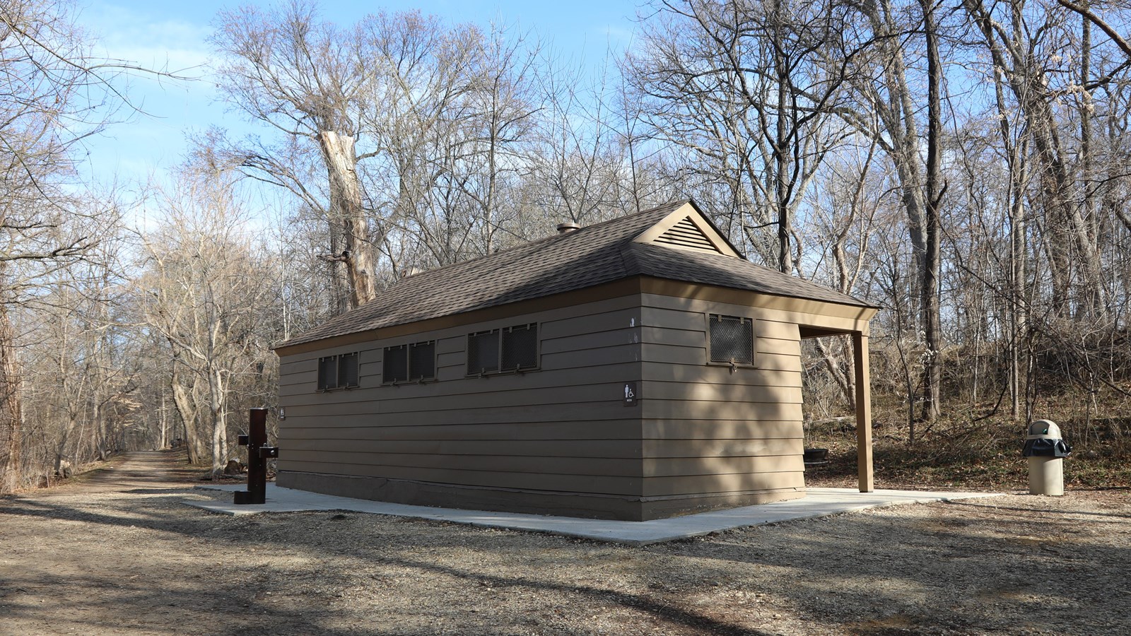 A single-story rectangular building sits on a concrete pad surrounded by a gravel path and trees.
