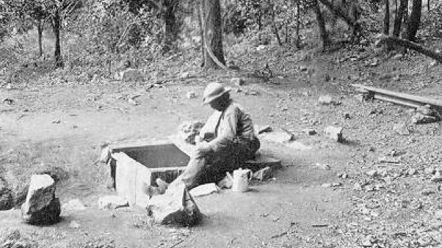 A man sits in a clearing next to a square opening containing a spring.
