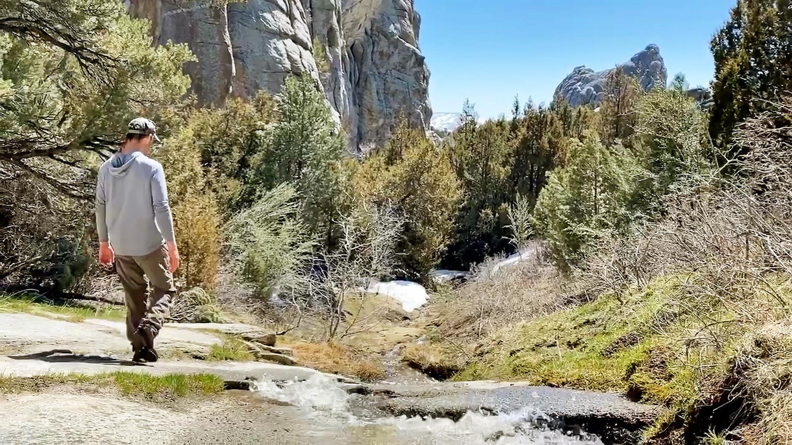A man hikes beside a creek among towering granites spires, pine, and juniper trees.