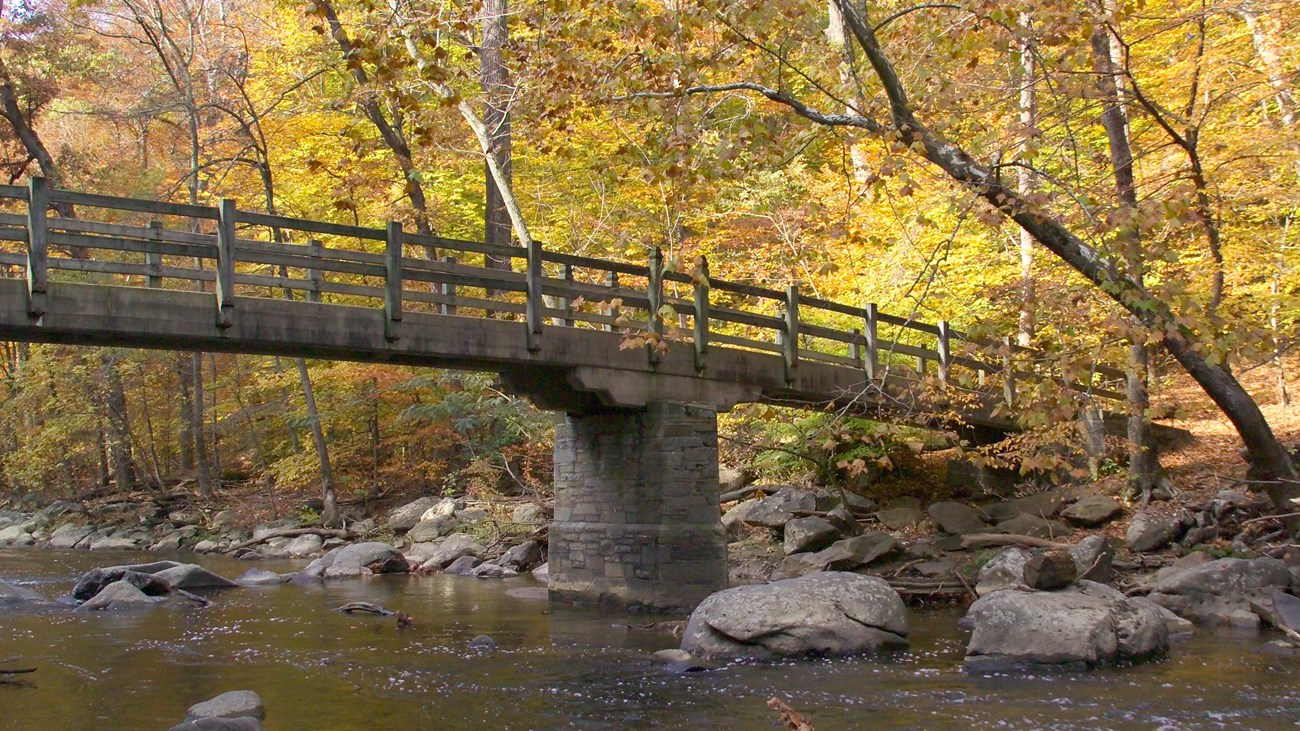 A wood and concrete bridge crosses a rocky creek with yellow and green trees on the far bank