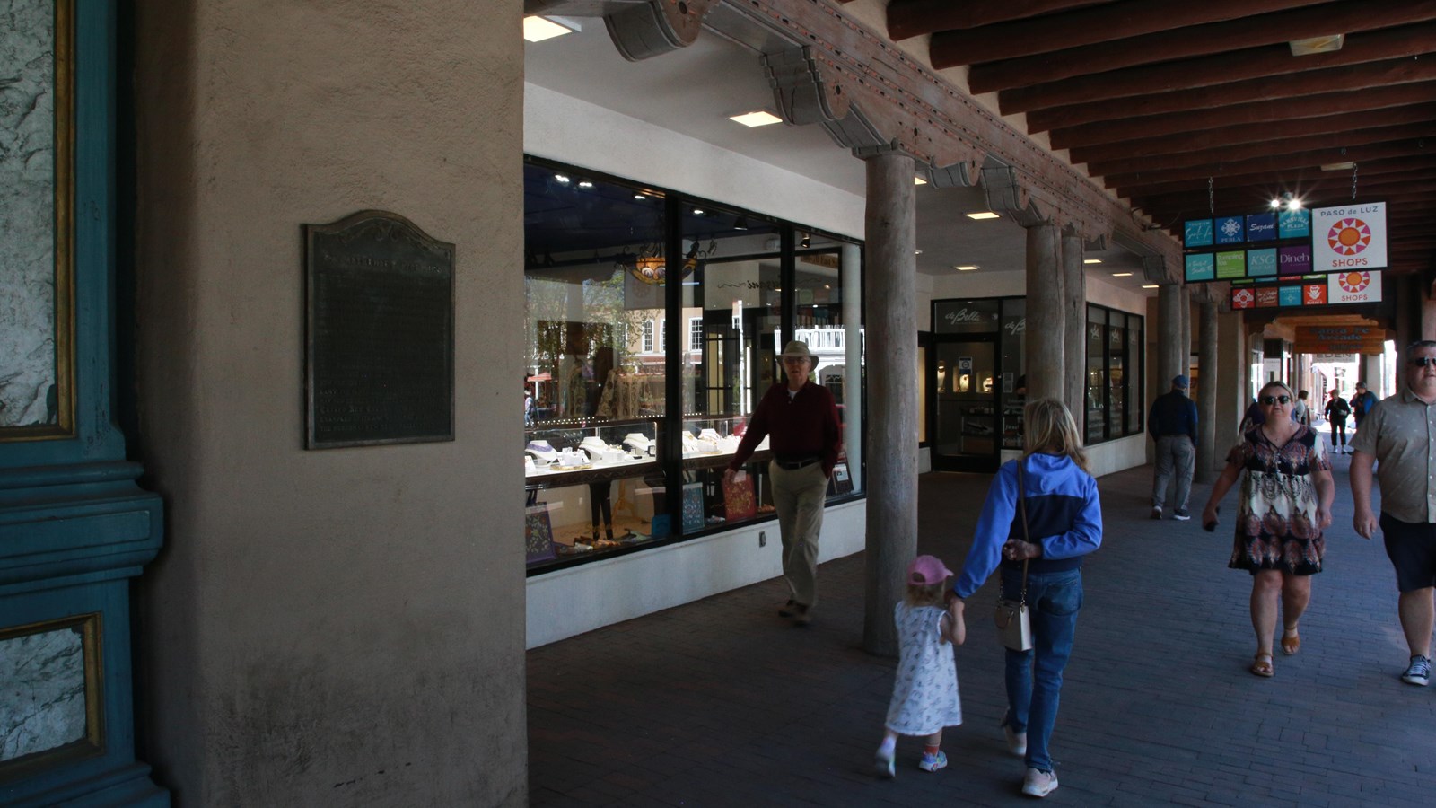 An aged bronze plaque is on an adobe wall in a line of business fronts covered by a veranda.