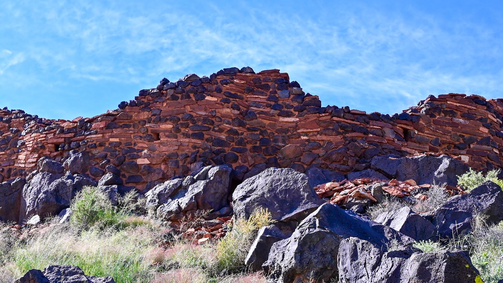 Looking up a grassy hill at a partial red sandstone and black basalt Pueblo with a blue sky. 