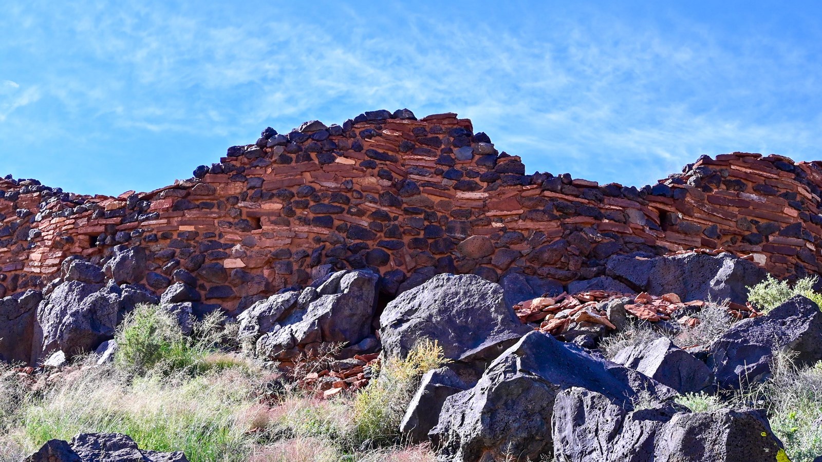 Looking up a grassy hill at a partial red sandstone and black basalt Pueblo with a blue sky.