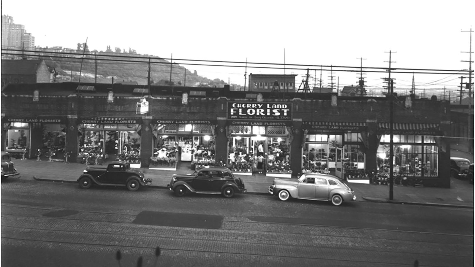 Depicts a street scene from an earlier era, featuring the Cherry Land Florist shop and vintage cars.