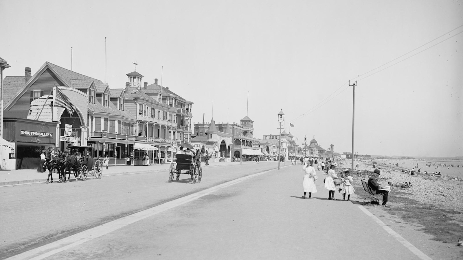 Ealry 20th century photo of people walking along a boardwalk with beach on the right. 