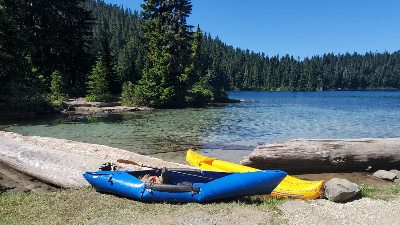A ten-foot long blue kayak sits on the bank of a lake in front of an inflatable yellow kayak.