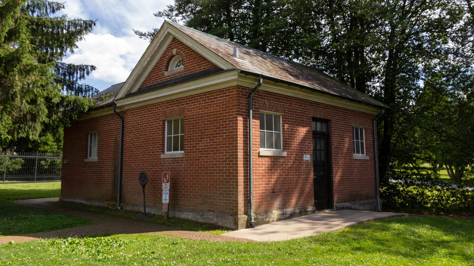 A small red brick structure with pathway through woods surrounding building.