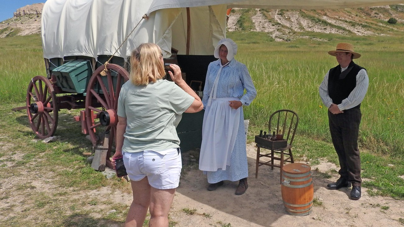 Two rangers, dressed as emigrants on the Oregon Trail, talk with a visitor next to a covered wagon.